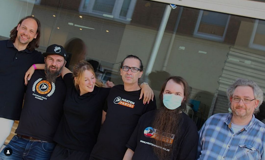 Es stehen 6 Personen vor einer Fensterscheibe. Die meisten tragen schwarze T-shirts mit hellem Piratenshlogo. Es in P Form geschwungenes Segel mit  Blau Weiß Roter Farbgebung in anspielung an die Schleswigholsteinflagge