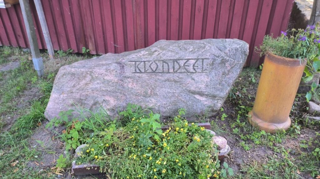 Stein mit eisernem Schriftzug "Klöndeel" und Blumen drumherum